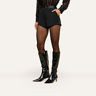 A person wearing the Colt Tuxedo Short crafted from suiting fabric, a sheer black long-sleeve top, sheer black tights, and shiny black knee-high boots stands against a plain white background.