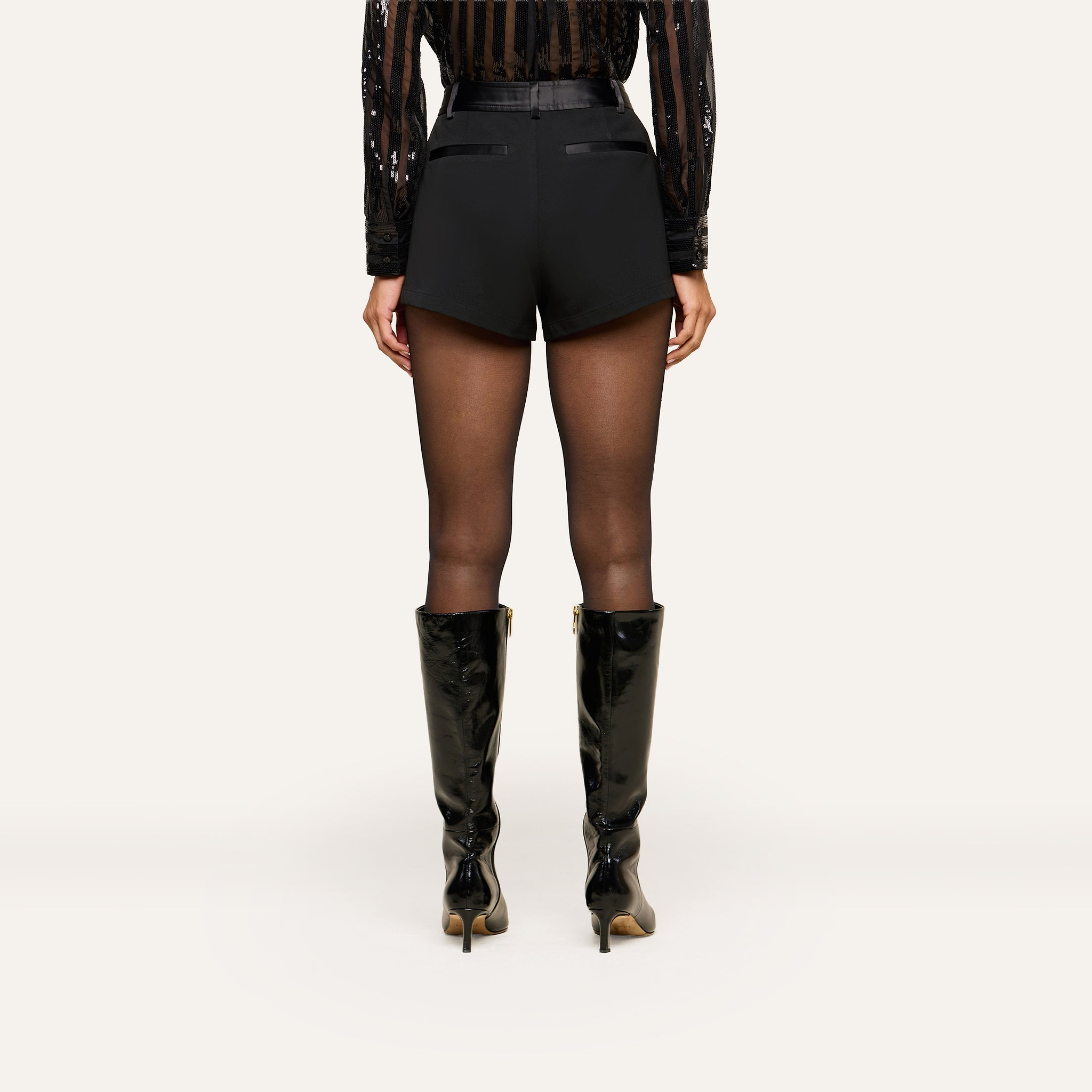 A person stands facing away, wearing a sheer black blouse, the Colt Tuxedo Short crafted from suiting fabric, sheer black tights, and black knee-high heeled boots against a plain light background.