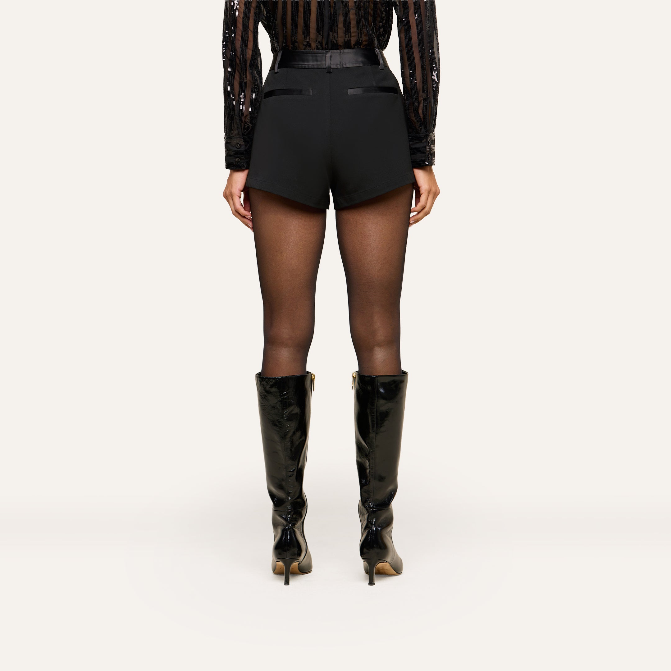A person stands facing away, wearing a sheer black blouse, the Colt Tuxedo Short crafted from suiting fabric, sheer black tights, and black knee-high heeled boots against a plain light background.