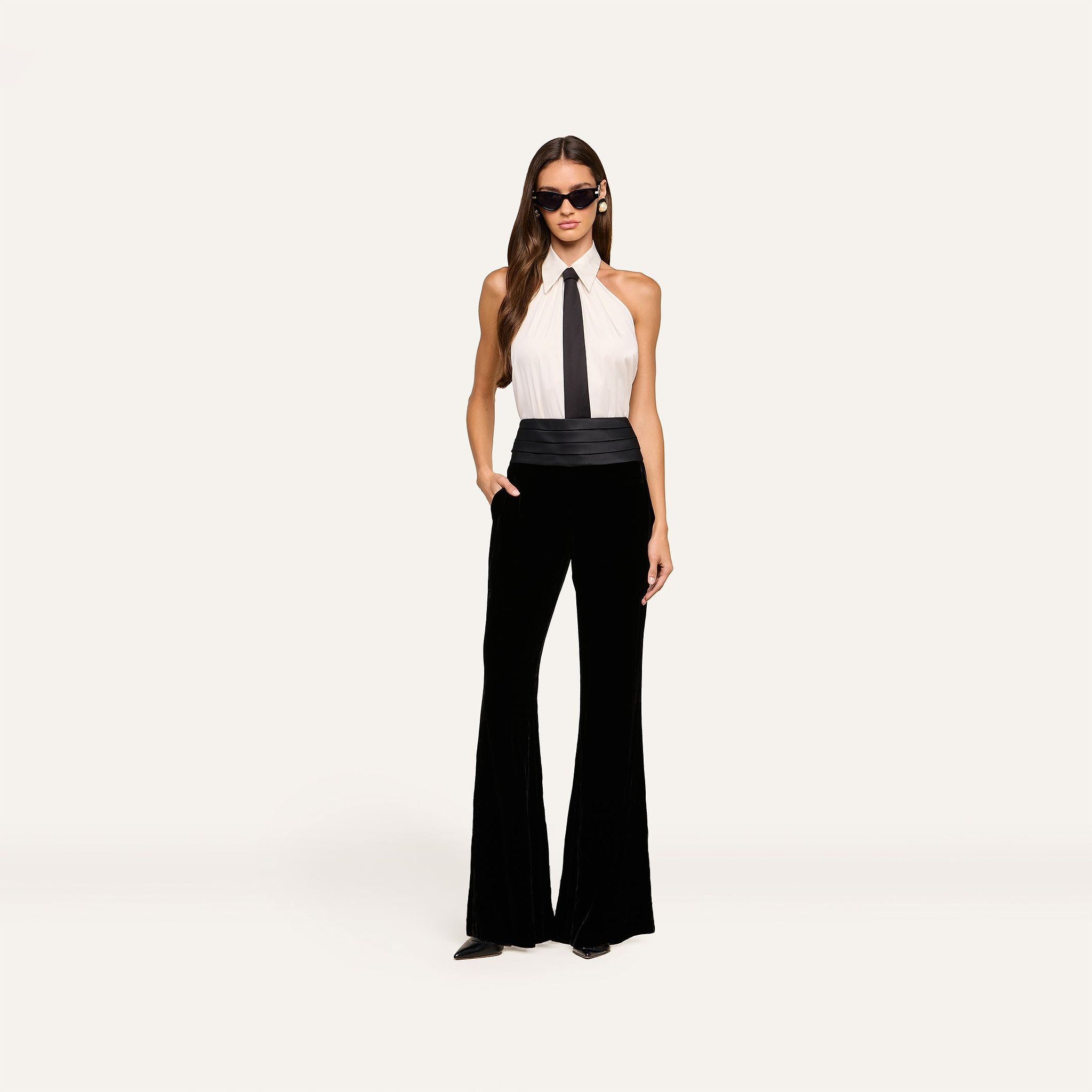 A woman in sunglasses wears a sleeveless white halter top with a black tie, the Prudence Cummerbund Trouser—high rise tuxedo-style black flared pants—and black heels, standing against a plain light background.