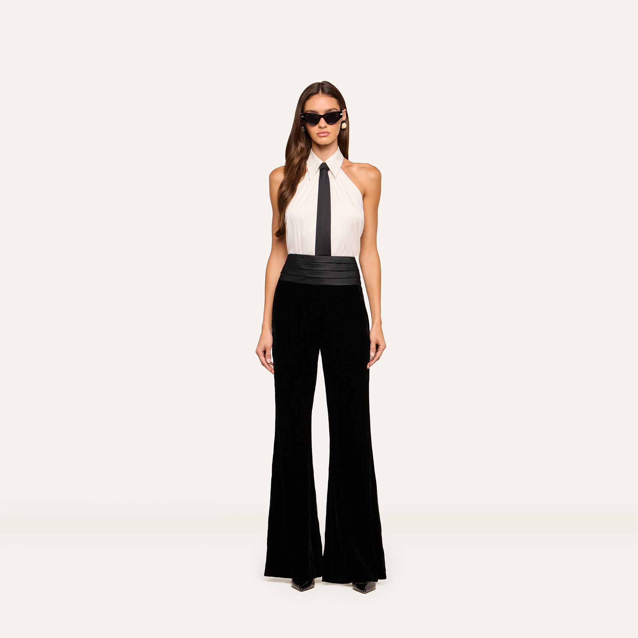A woman wearing sunglasses, a sleeveless white halter top with a black necktie, a black cummerbund, and the Prudence Cummerbund Trouser—high rise, wide-leg pants in tuxedo style—stands against a neutral background.