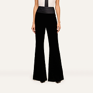 A woman wearing the Prudence Cummerbund Trouser, a pair of high rise velvet trousers, and a sleeveless white top with a black vertical stripe stands against a plain light background.