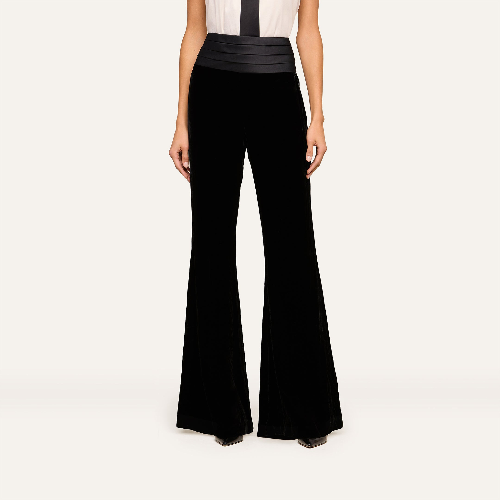 A woman wearing the Prudence Cummerbund Trouser, a pair of high rise velvet trousers, and a sleeveless white top with a black vertical stripe stands against a plain light background.
