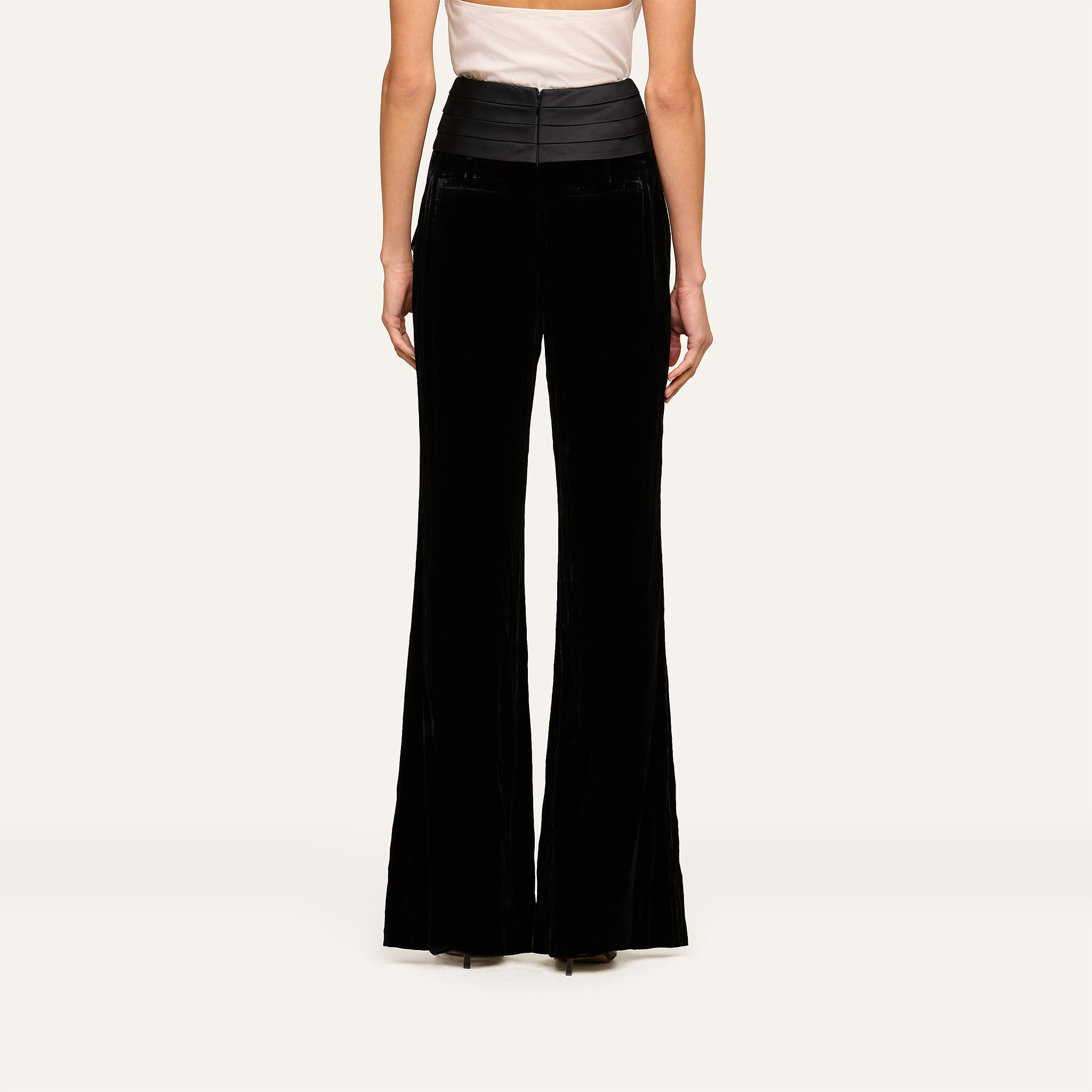 A person is shown from the back against a plain light background, wearing the Prudence Cummerbund Trouser, which features a high-rise fit, black velvet fabric, and wide legs, paired with a sleeveless beige top.
