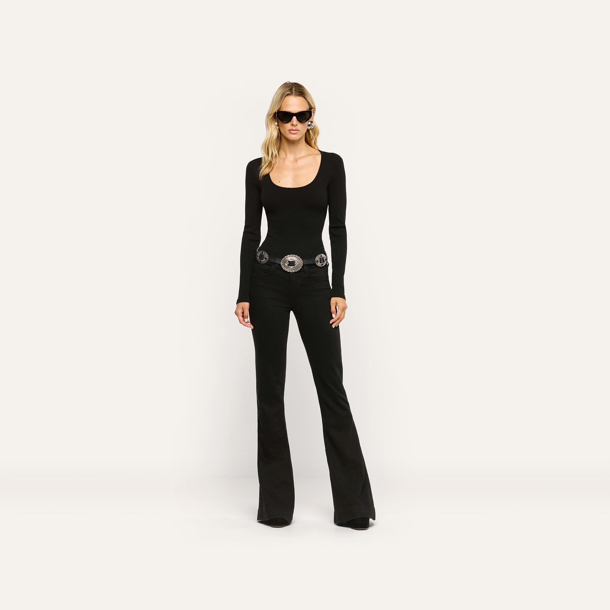 A woman wearing a black long-sleeved scoop-neck top, a silver-accented belt, and Her Story Flare Jean.