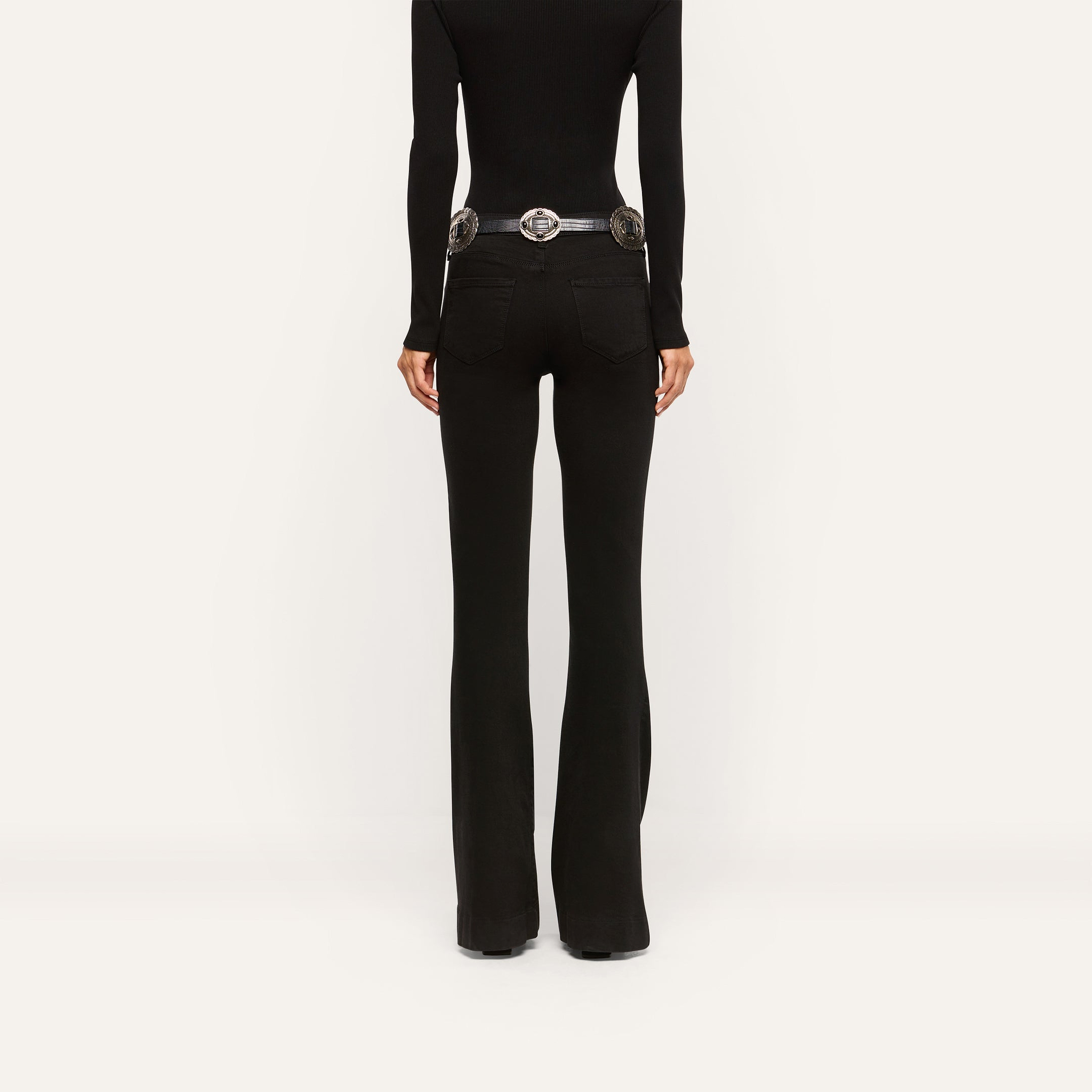 Back view of black slim-cut Her Story Flare Jean worn with a black long-sleeve top and a silver-buckled belt.