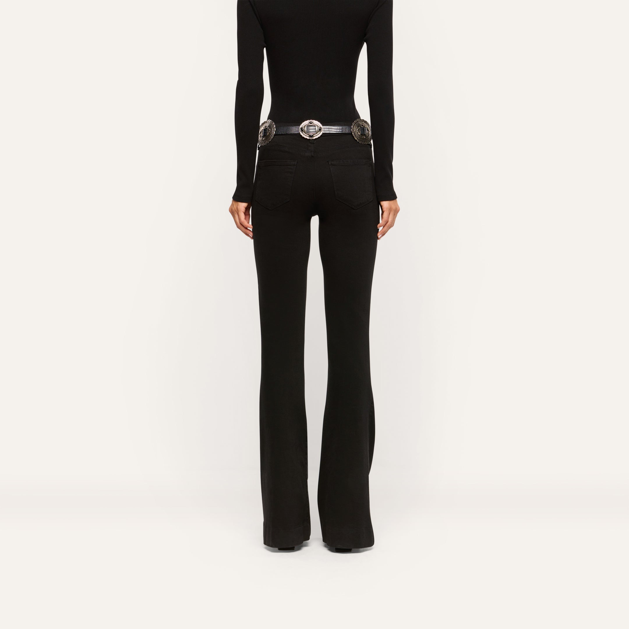 Back view of black slim-cut Her Story Flare Jean worn with a black long-sleeve top and a silver-buckled belt.