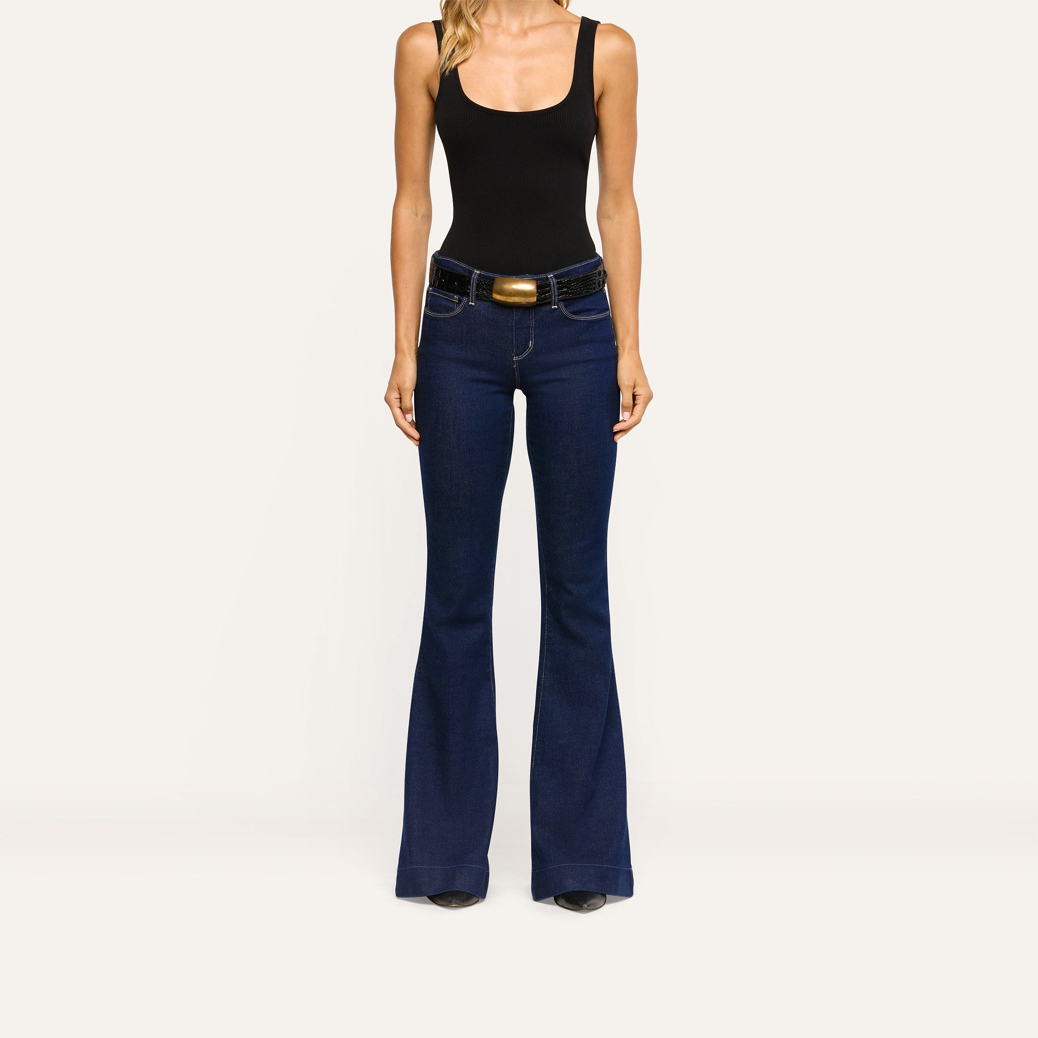 Her Story Flare Jean in slim-cut dark indigo, worn with a black tank top and a black belt with a gold buckle.