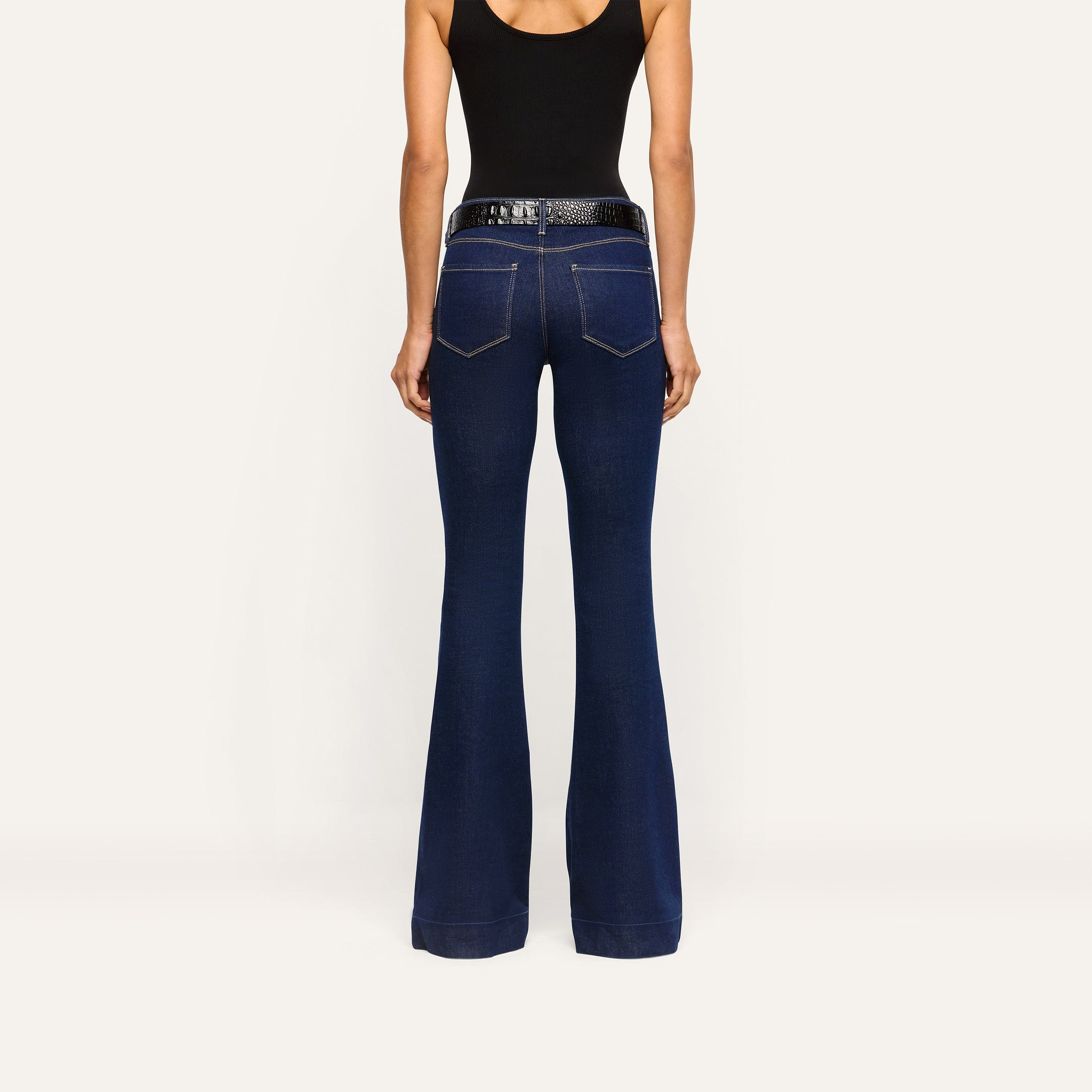 Back view of Her Story Flare Jean in slim-cut dark indigo, worn with a black tank top and a black textured belt.