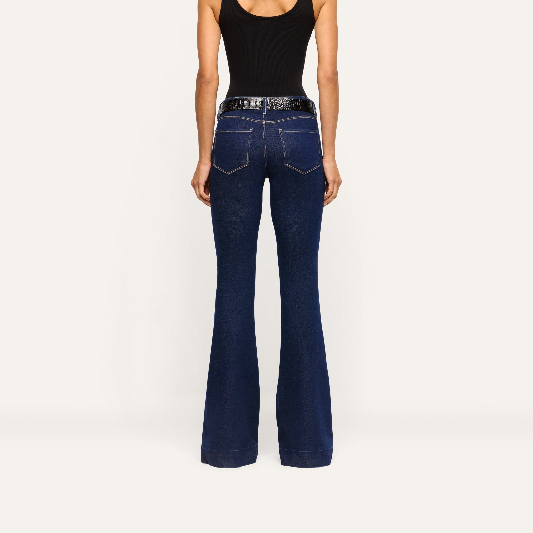 Back view of Her Story Flare Jean in slim-cut dark indigo, worn with a black tank top and a black textured belt.