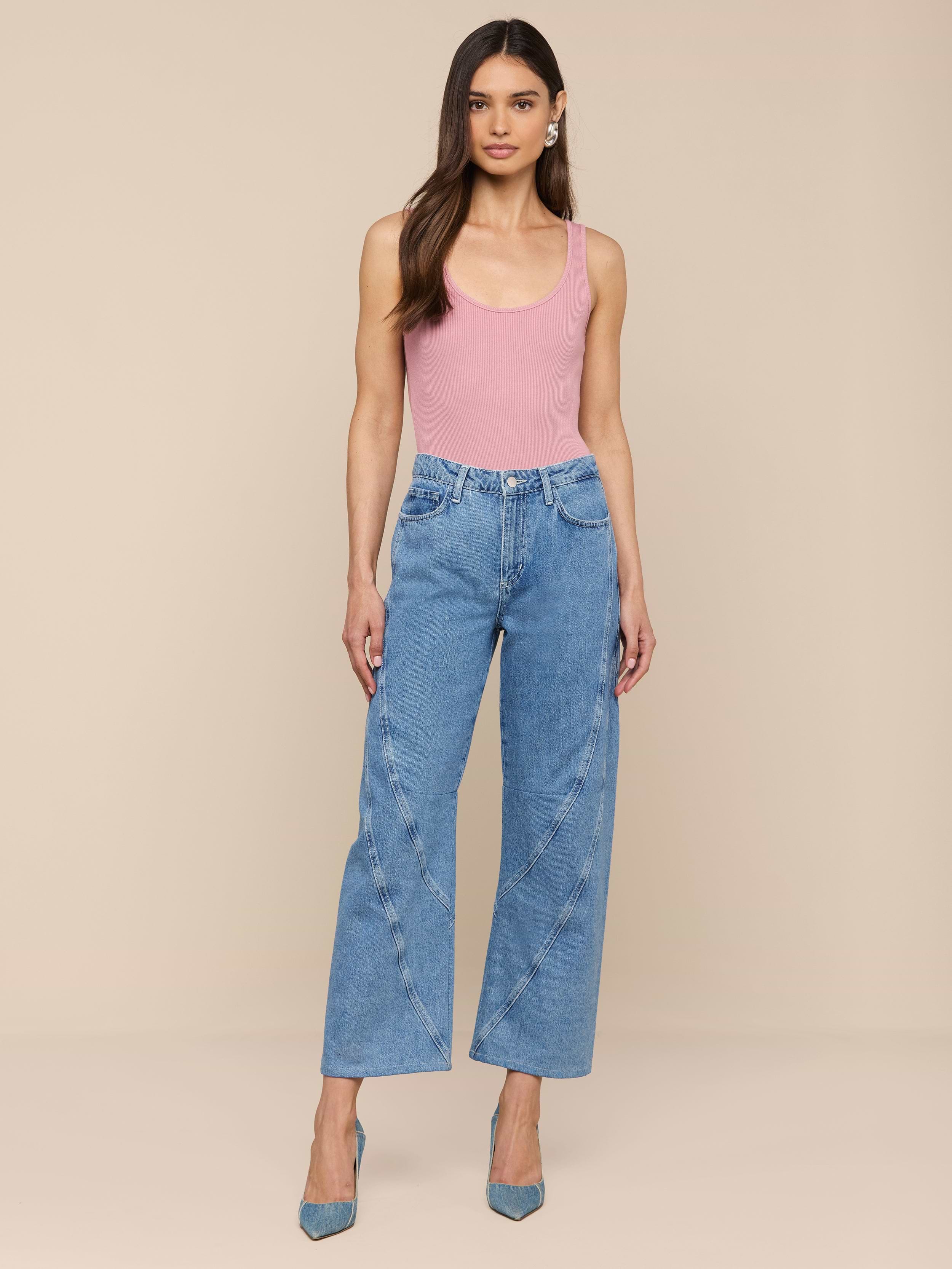 Une femme se tient debout sur un fond beige, vêtue dun débardeur rose, de talons bleus à bouts pointus et du jean Frasier Rodeo Jean - taille haute, coupe slouchy, tout en coton avec des coutures incurvées. Ses longs cheveux tombent librement et ses bras sont détendus le long de son corps.