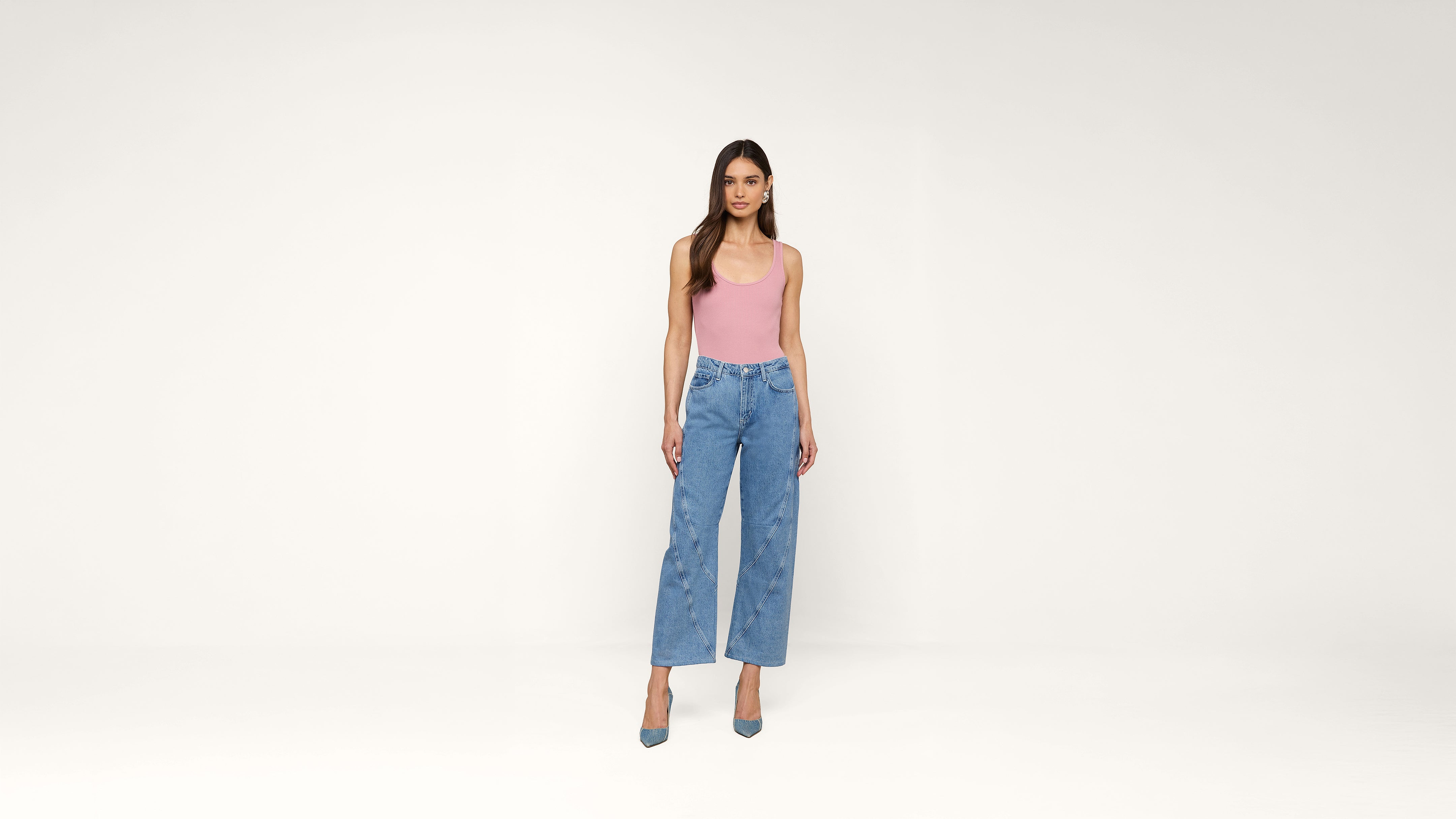 A woman stands against a plain white background, wearing a pink sleeveless top, blue high-waisted jeans, and blue heels. She has long, loose hair and a neutral expression.