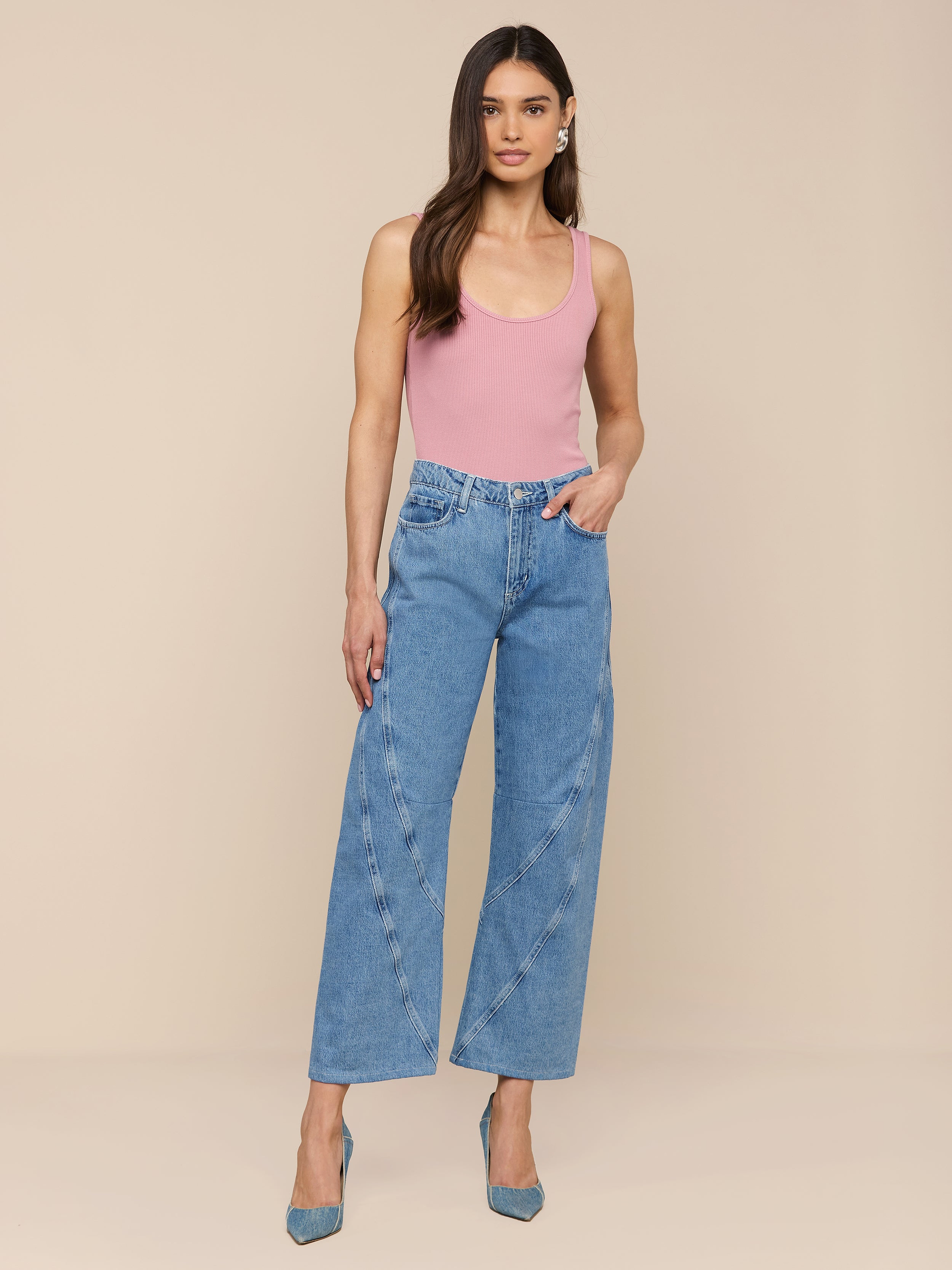 Une femme aux longs cheveux bruns porte un haut rose sans manches, un jean Frasier Rodeo en denim tout coton avec des coutures incurvées, et des talons bleus, sur un fond beige uni.