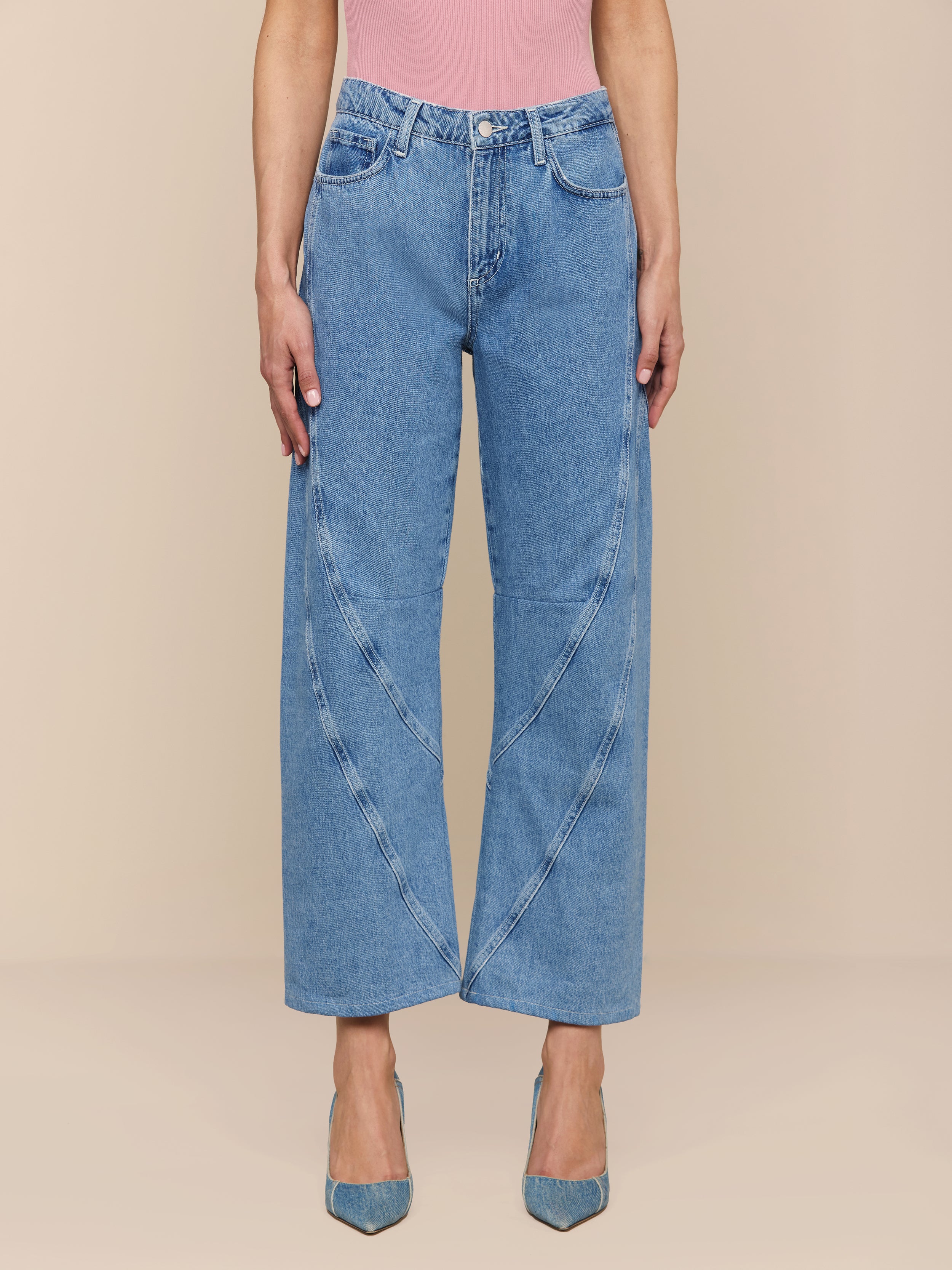 Portant le Frasier Rodeo Jean - un jean bleu à taille haute et à jambe large avec des coutures incurvées sur le devant - une personne lassocie à un haut rose et à des talons bleus à bouts pointus, sur un fond beige uni.