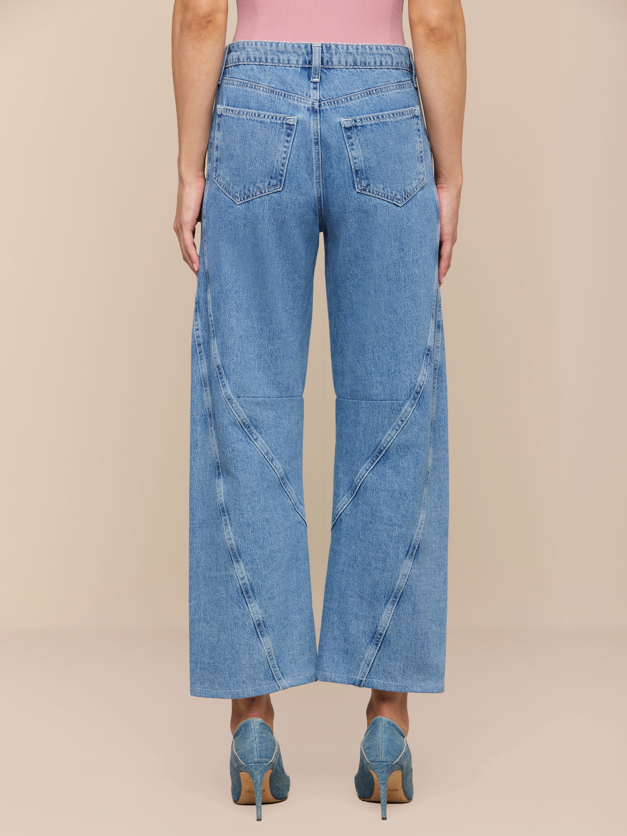 Modèle portant le Frasier Rodeo Jean à jambe large, coupe slouchy, en denim tout coton avec coutures incurvées et poches arrière, associé à des talons sarcelle et un top rose, montré de dos sur un fond beige uni.