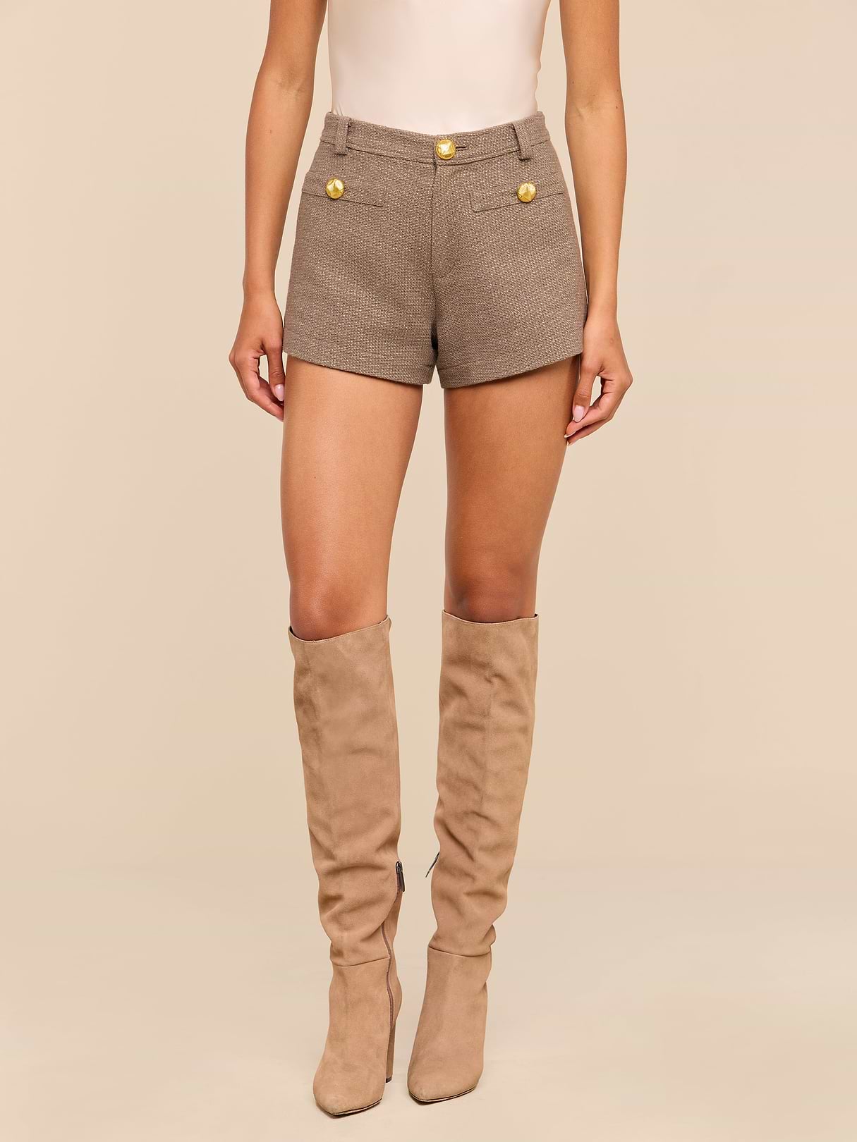 A person wearing the Dionne Tweed Short—high rise brown shorts with gold buttons made from luxurious Italian Lurex tweed—paired with taupe knee-high boots, stands against a beige background. Only the body from shoulders to feet is visible.