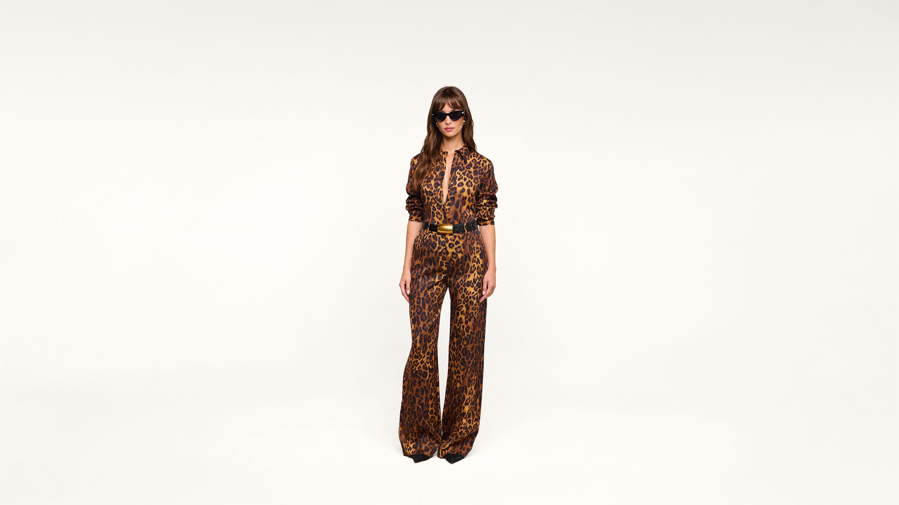 A woman wearing sunglasses and a leopard print jumpsuit stands against a plain white background, facing forward with her hands by her sides.