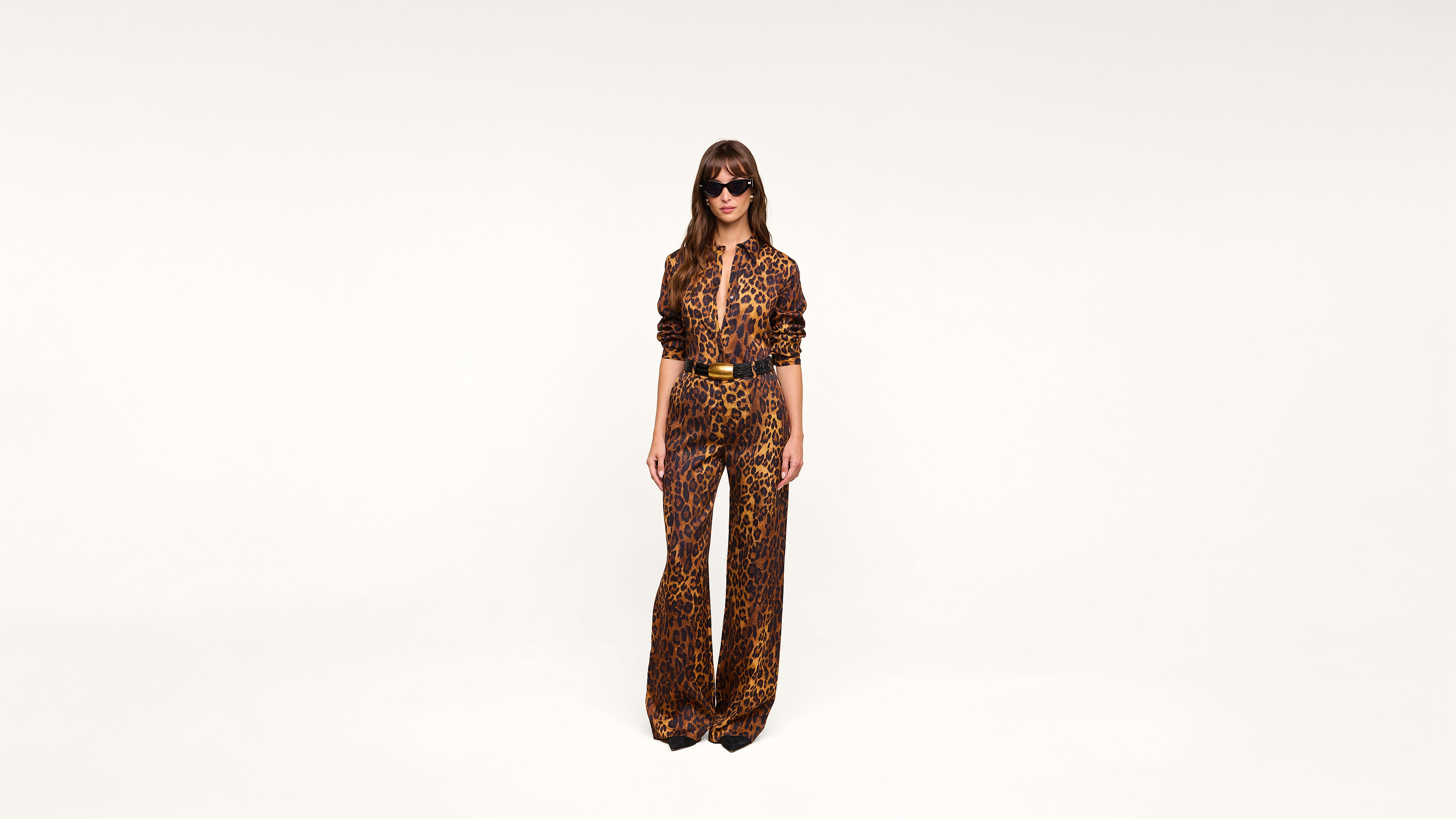 A woman wearing sunglasses and a leopard print jumpsuit stands against a plain white background, facing forward with her hands by her sides.