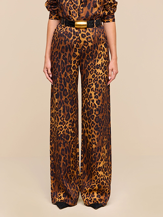A person wearing the Livvy Trouser—tan leopard print wide-leg pants crafted from printed twill—with a matching top and a black belt featuring a large gold buckle, standing against a beige background.