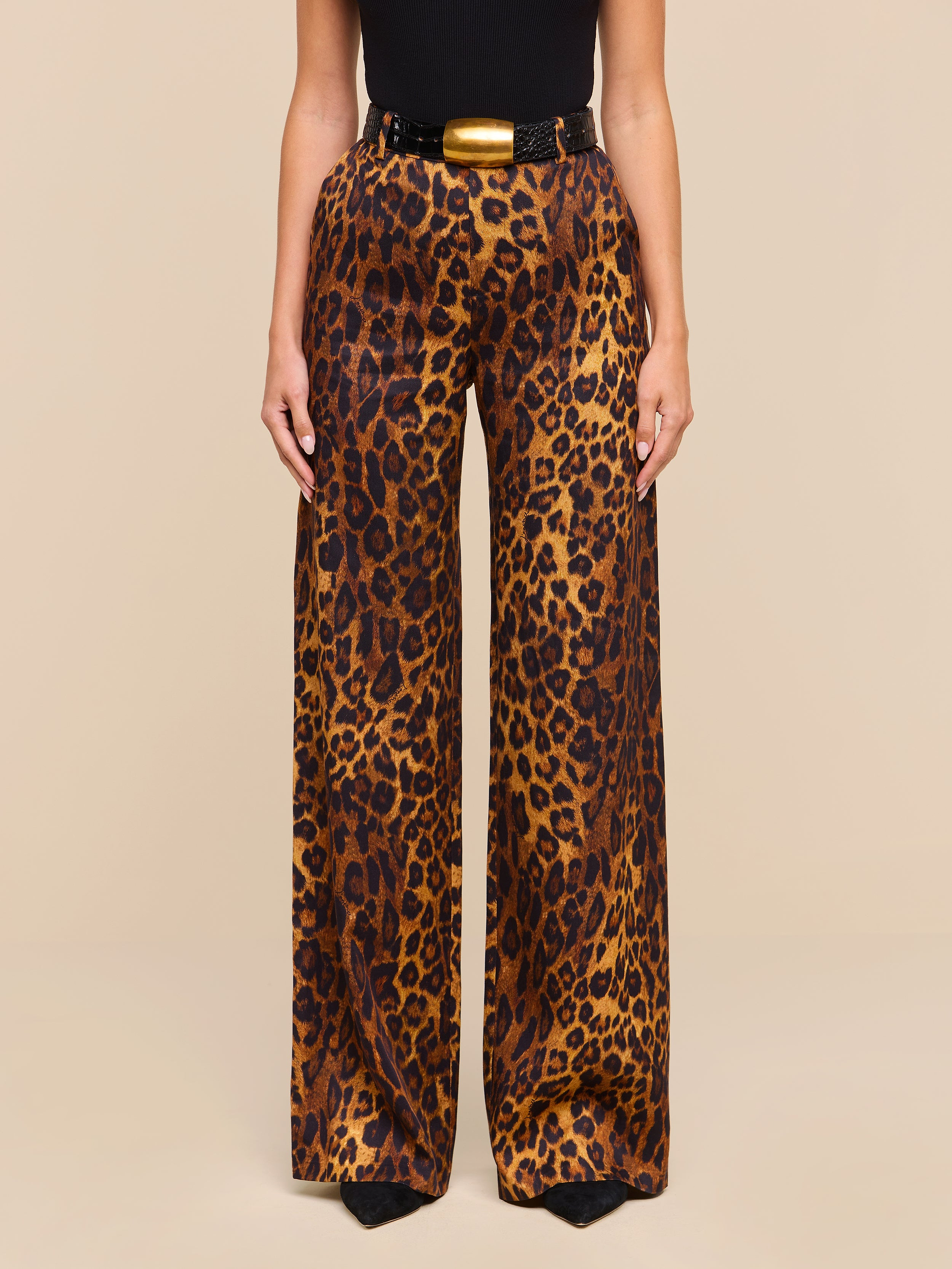 A person wearing the Livvy Trouser, featuring a high-waisted, wide-leg silhouette in tan leopard print, paired with a black belt and a fitted black top, stands against a plain white background.