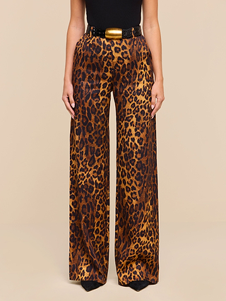 A person wearing the Livvy Trouser, a high-waisted, straight-leg pair in a bold tan leopard print, styled with a black belt featuring a large gold buckle and a sleek black top, stands against a plain beige background.
