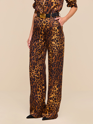 Wearing the Livvy Trouser, a pair of high-waisted, wide-leg tan leopard print pants crafted from printed twill, paired with a matching long-sleeve top, black belt with a gold buckle, and pointed black shoes, this person stands with one hand in their pocket against a beige background.