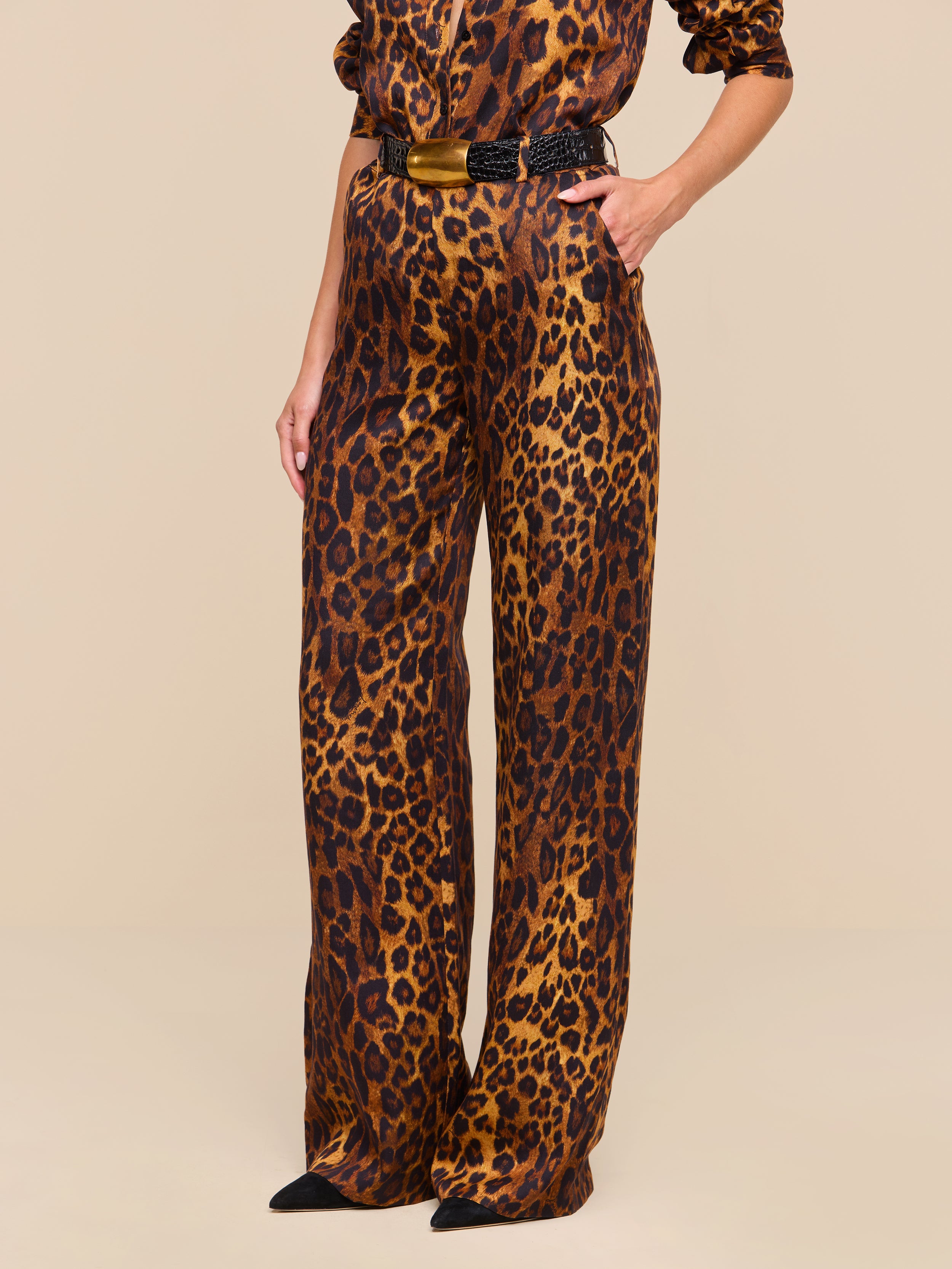 A person is shown wearing the Livvy Trouser, a tan leopard print jumpsuit featuring wide, straight-leg trousers and a black belt, standing against a plain white background with their face not visible.