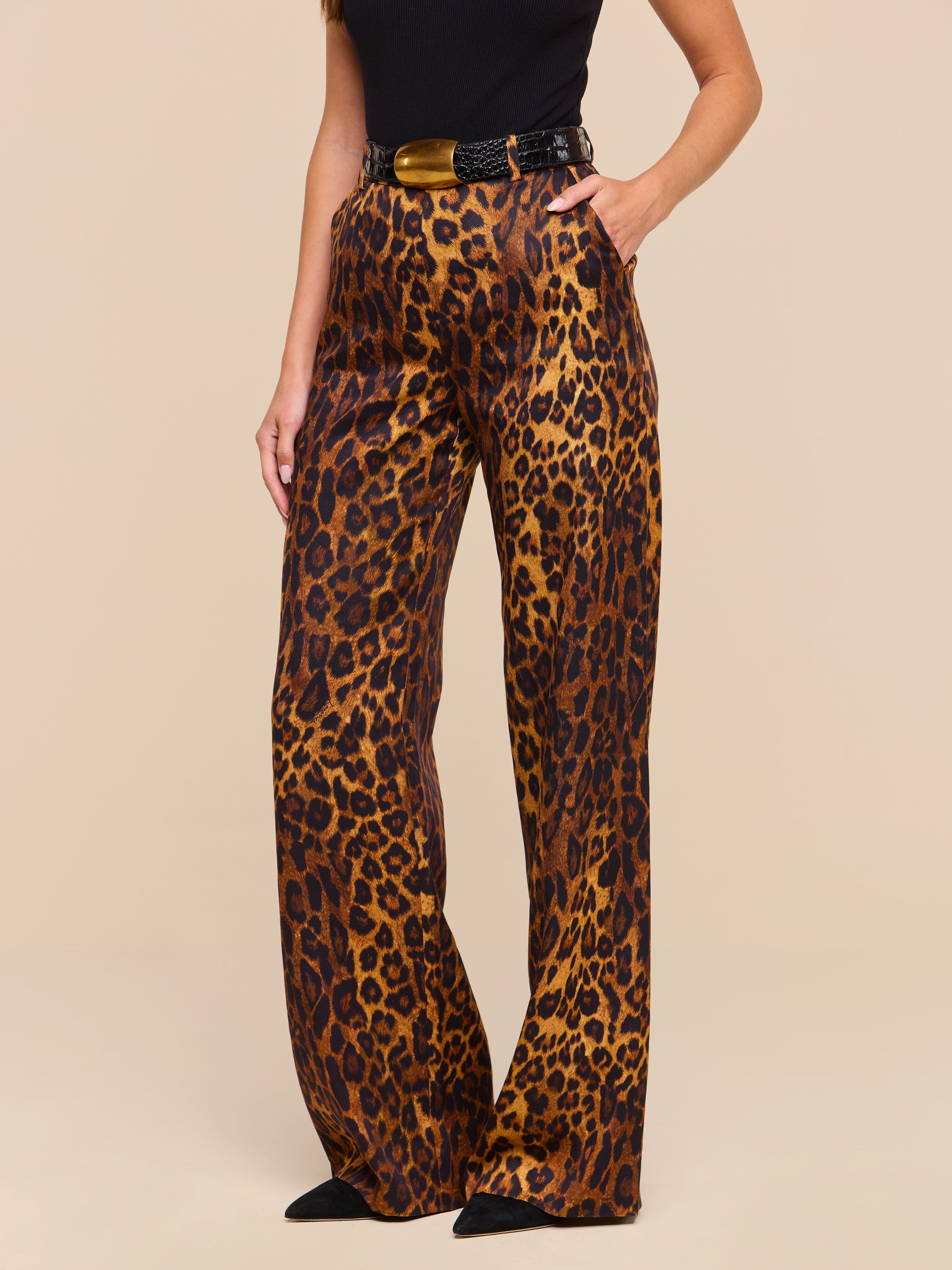 A woman is shown from torso to feet wearing the Livvy Trouser, which features a high-waisted, wide-leg silhouette in tan leopard print, paired with a black top and pointed black shoes against a plain background.