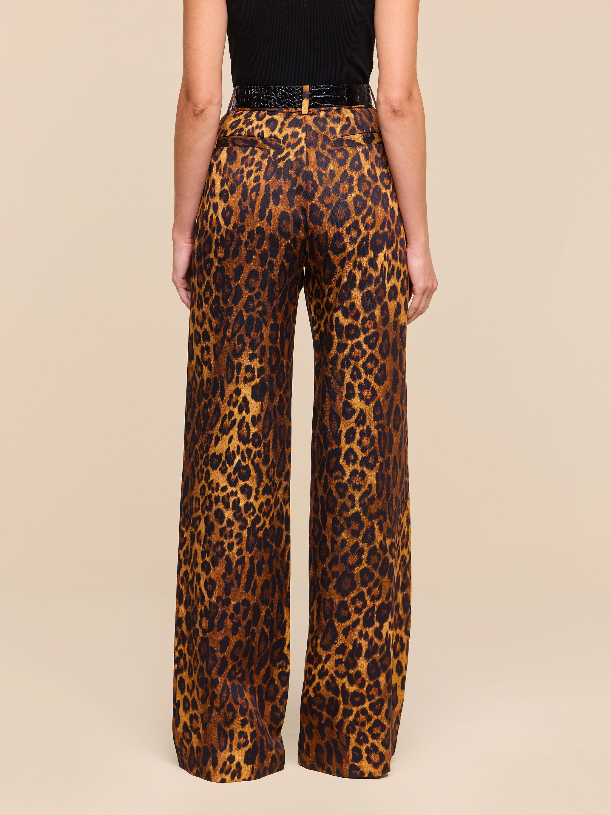 A person stands with their back to the camera, wearing the Livvy Trouser—high-waisted, straight-leg tan leopard print pants—paired with a black top against a plain white background.