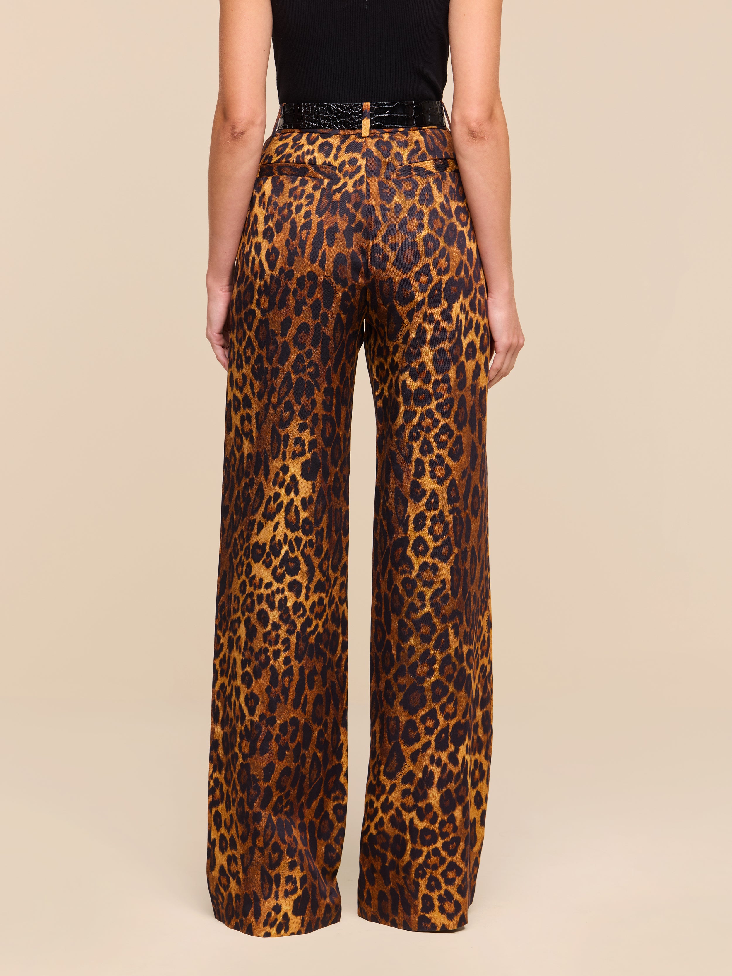 A person stands with their back to the camera, wearing the Livvy Trouser—high-waisted, straight-leg tan leopard print pants—paired with a black top against a plain white background.