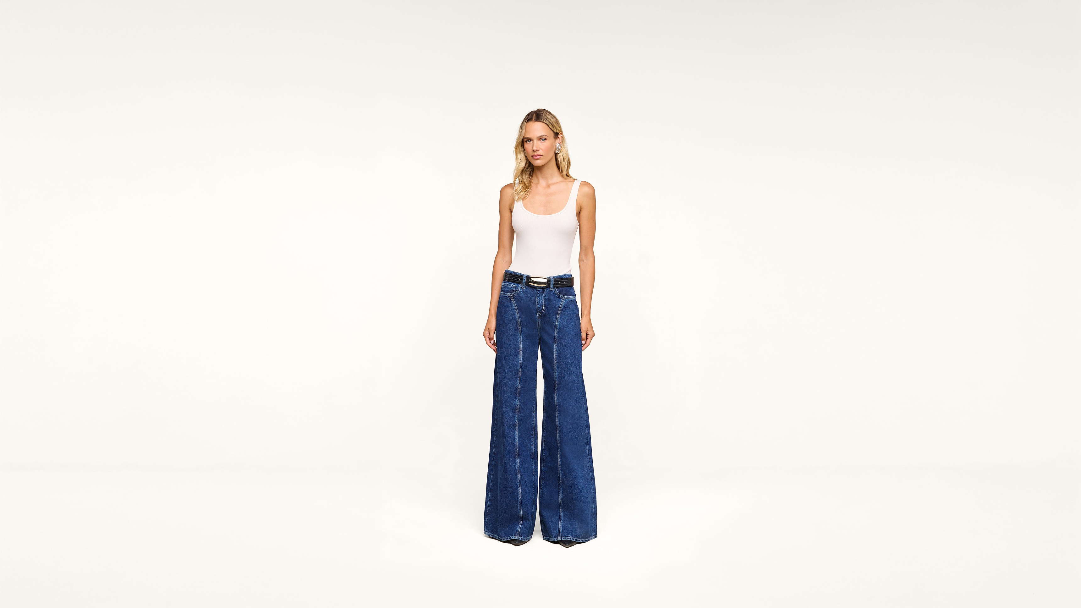 A woman stands against a plain white background wearing a white tank top, blue high-waisted wide-leg jeans, and a black belt.