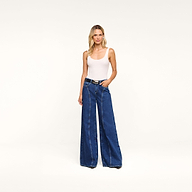 A woman stands against a plain background, wearing a white sleeveless top and the Lestat Wide-Leg Jean, high-rise jeans crafted from all-cotton denim. She has long, wavy blonde hair and her hands are in her pockets.