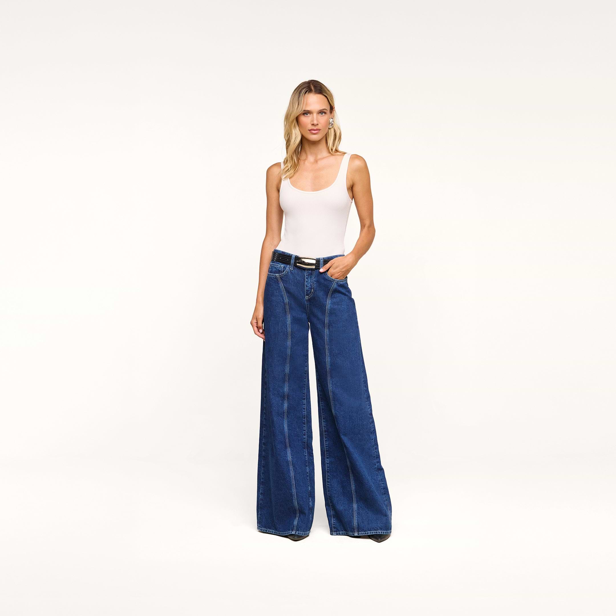 A woman stands against a plain background, wearing a white sleeveless top and the Lestat Wide-Leg Jean, high-rise jeans crafted from all-cotton denim. She has long, wavy blonde hair and her hands are in her pockets.