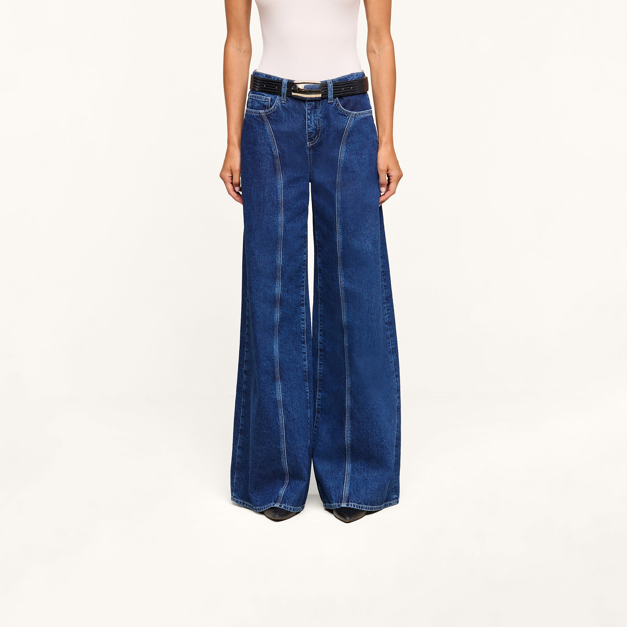 A model, shown from the shoulders down and standing against a plain white background, wears the Lestat Wide-Leg Jean—a high-rise, wide-leg style crafted from all-cotton denim—paired with a white sleeveless top and black belt.