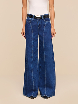 A person wearing a white sleeveless top, the Lestat Wide-Leg Jean crafted from all-cotton denim with contrast stitching, a black belt, and black shoes, standing against a beige background.