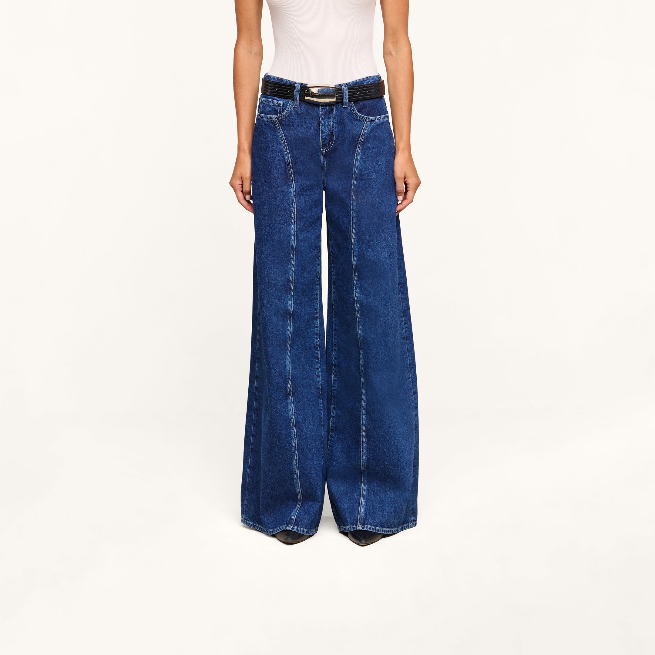A model, shown from the shoulders down and standing against a plain white background, wears the Lestat Wide-Leg Jean—a high-rise, wide-leg style crafted from all-cotton denim—paired with a white sleeveless top and black belt.