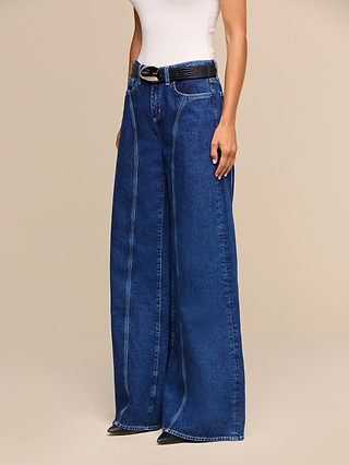 A person wearing a white sleeveless top, a black belt, black pointed shoes, and the Lestat Wide-Leg Jean—a pair of high-rise, all-cotton denim jeans with prominent seams—stands against a beige background.