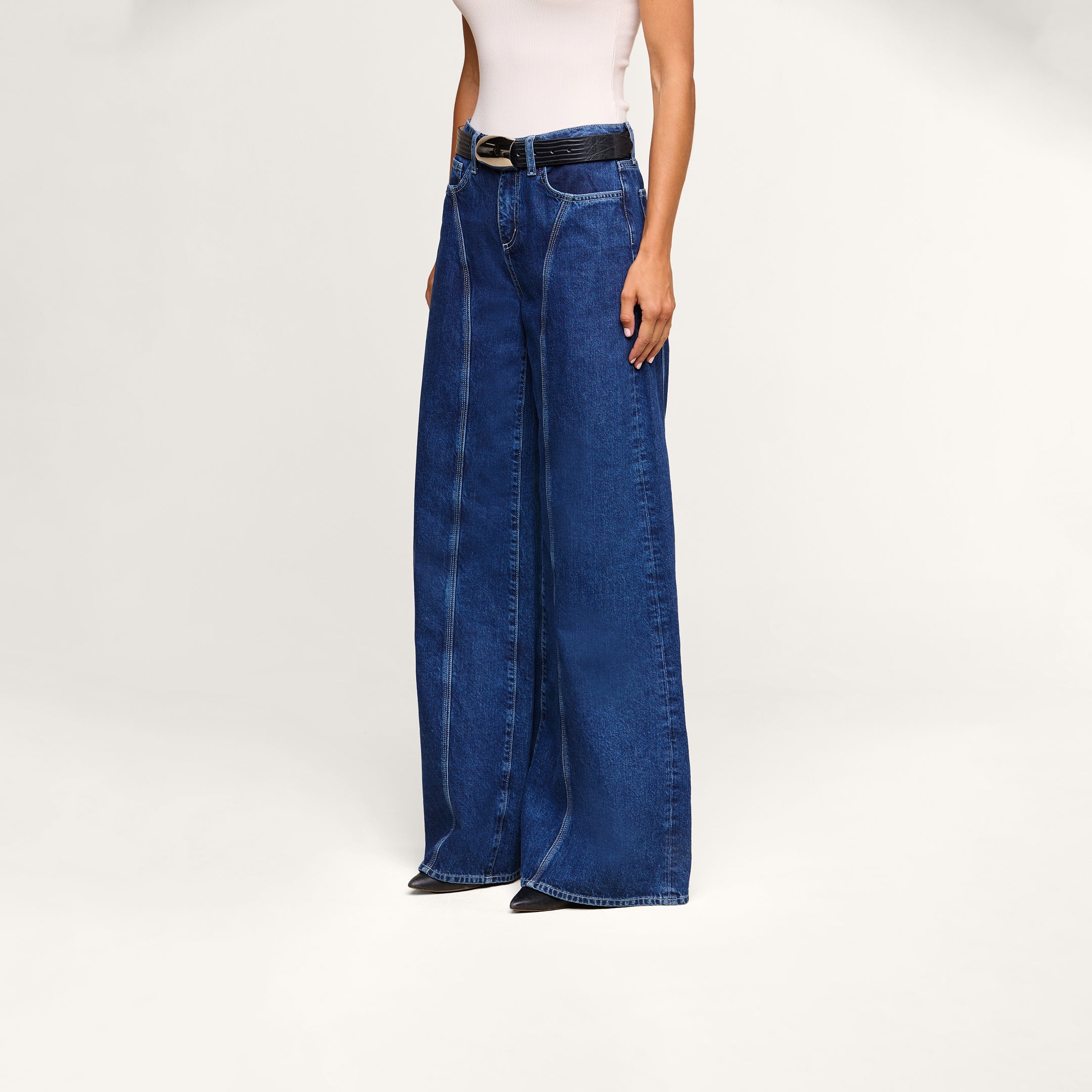 A person wearing the Lestat Wide-Leg Jean—a high-rise, wide-leg style made from all-cotton blue denim—along with a sleeveless white top and a black belt stands against a plain light background. Their face is not visible in the image.