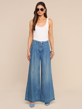 A woman stands against a beige background wearing sunglasses, a white tank top, and the Jayani Pleated Wide-Leg Jean crafted from non-stretch denim. She pairs this look with blue heels, has long brown hair, and wears bracelets on her right wrist.