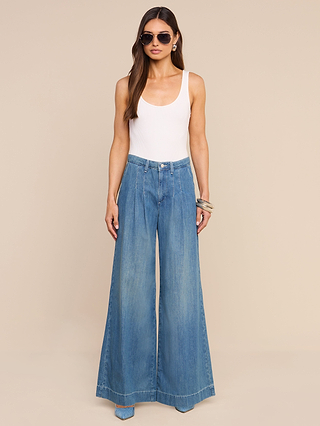 A woman stands against a beige background wearing sunglasses, a white sleeveless top, and the Jayani Pleated Wide-Leg Jean crafted from lightweight blue denim, accessorized with bracelets.