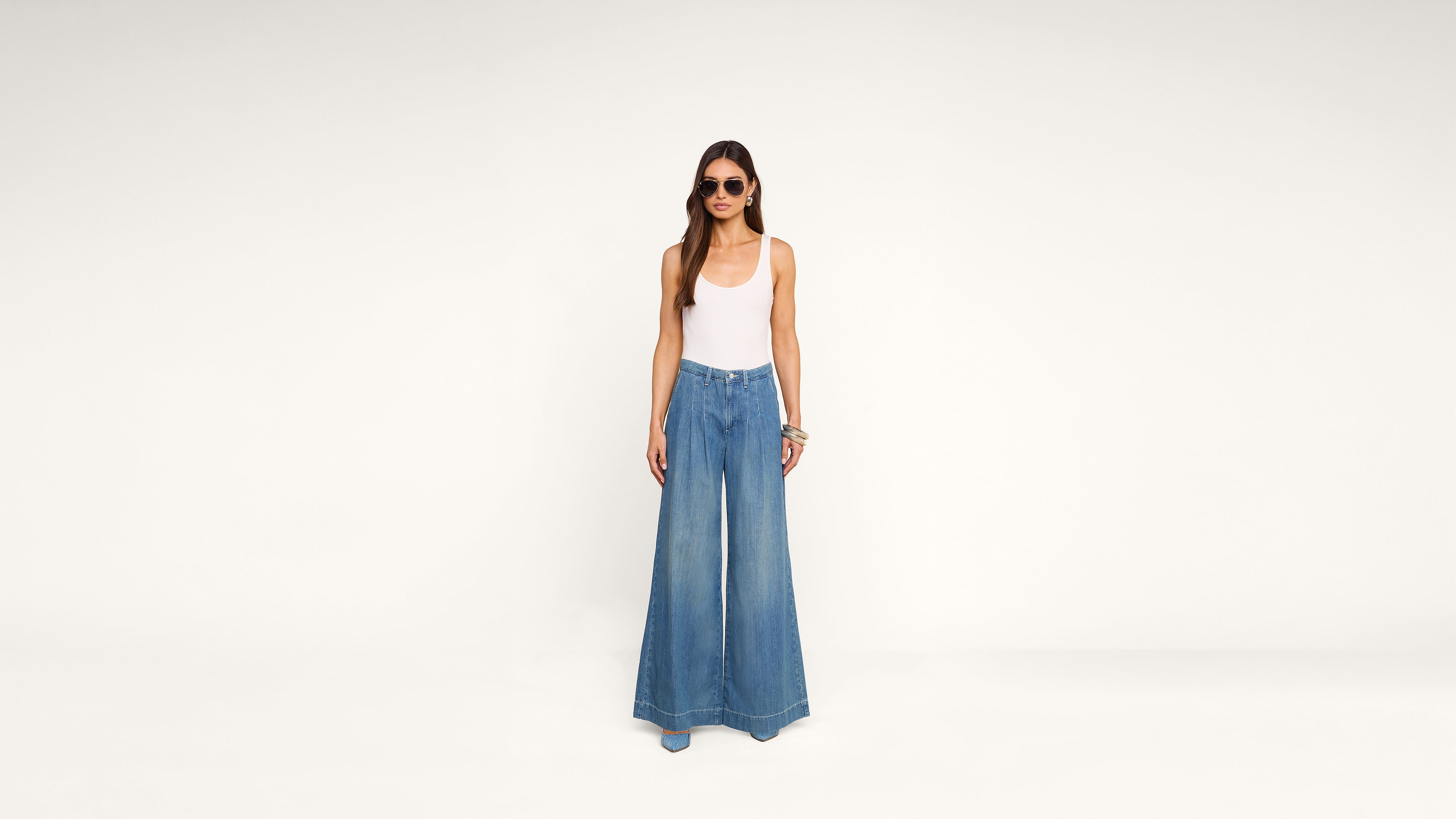 A woman wearing sunglasses, a white tank top, and wide-leg blue jeans stands against a plain white background.