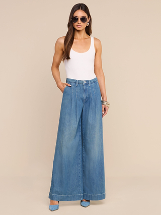 A woman wearing the Jayani Pleated Wide-Leg Jean—a lightweight, wide-leg blue jean—paired with a white sleeveless top, blue heels, sunglasses, and bracelets, stands with one hand in her pocket against a beige background.