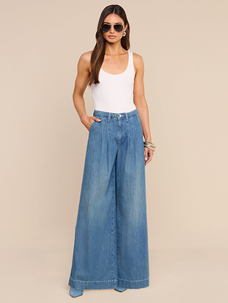 A woman wearing sunglasses, a white tank top, and the Jayani Pleated Wide-Leg Jean crafted from lightweight denim stands with one hand in her pocket against a beige background.