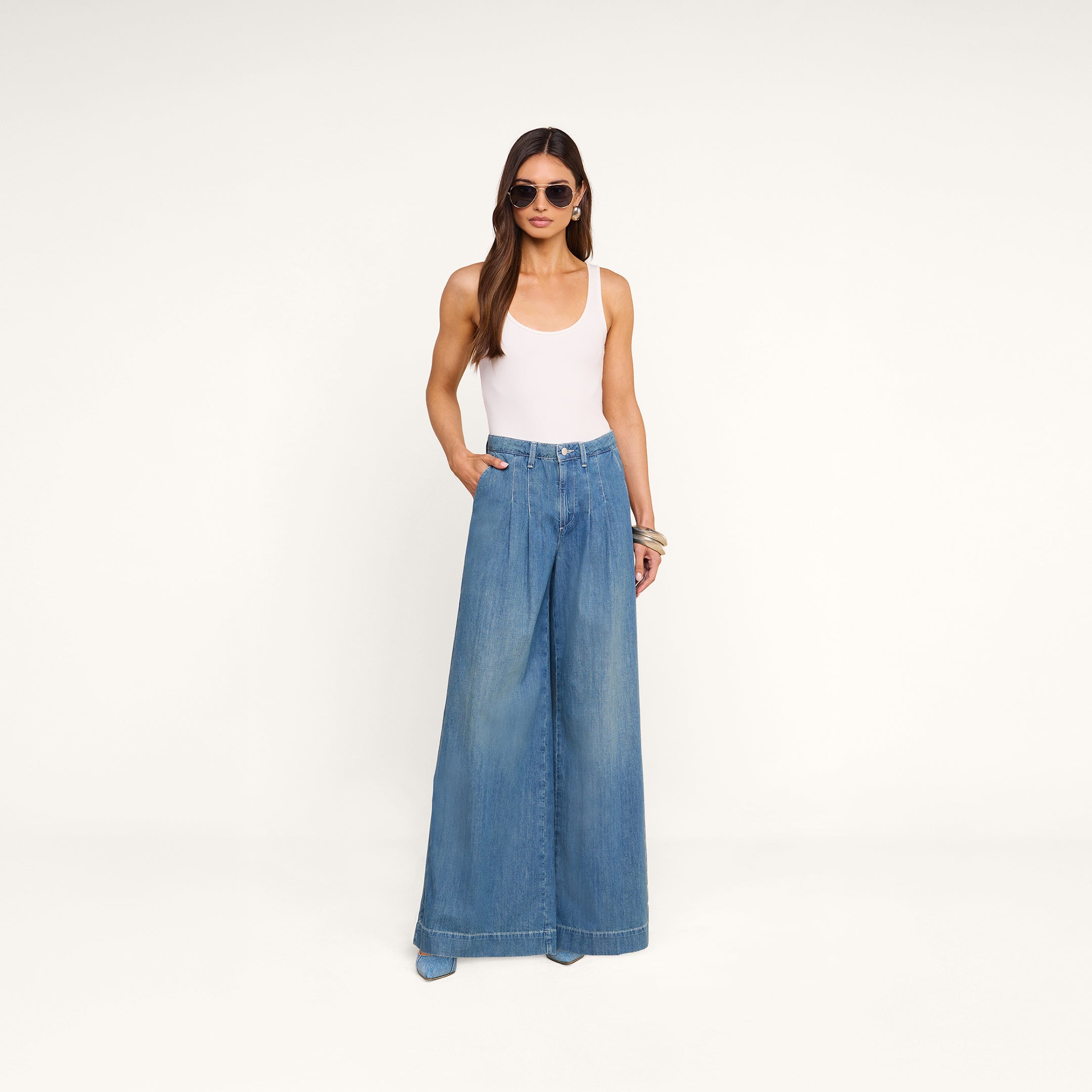 A woman wearing a white tank top, the Jayani Pleated Wide-Leg Jean in featherweight denim, sunglasses, and bracelets stands with her hands in her pockets against a plain white background.