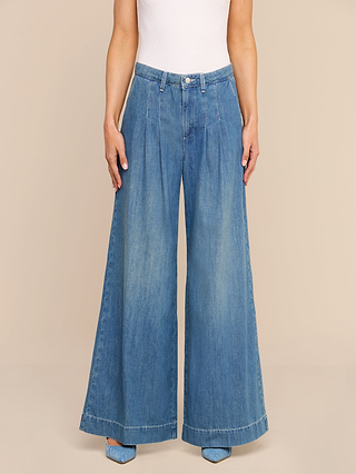 Wearing the Jayani Pleated Wide-Leg Jean in blue, crafted from non-stretch denim, and a white sleeveless top, this person stands against a beige background with their hands at their sides. Only the lower torso and legs are visible.
