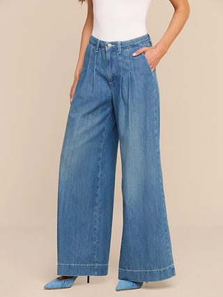 A person wearing a white sleeveless top, the Jayani Pleated Wide-Leg Jean—lightweight blue jeans with front pleats crafted from non-stretch denim—and pointed blue high heels stands against a beige background.
