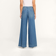 Una persona che indossa i jeans Jayani Pleated Wide-Leg, realizzati in denim non elasticizzato, con vita alta e gamba larga, di colore blu, abbinati a un top bianco senza maniche e ripresi di schiena su uno sfondo beige.