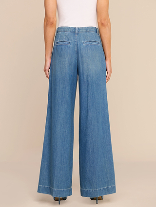A person wearing the Jayani Pleated Wide-Leg Jean, crafted from non-stretch denim in a high-waisted, wide-leg fit blue style, paired with a sleeveless white top and shown from the back against a beige background.
