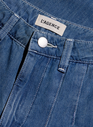 Close-up of the Jayani Pleated Wide-Leg Jean in blue denim, showcasing a white L’AGENCE label, a silver button, and prominent white stitching along the waistband and front seam. Crafted from lightweight denim for comfort, these jeans feature a chic high-rise silhouette.