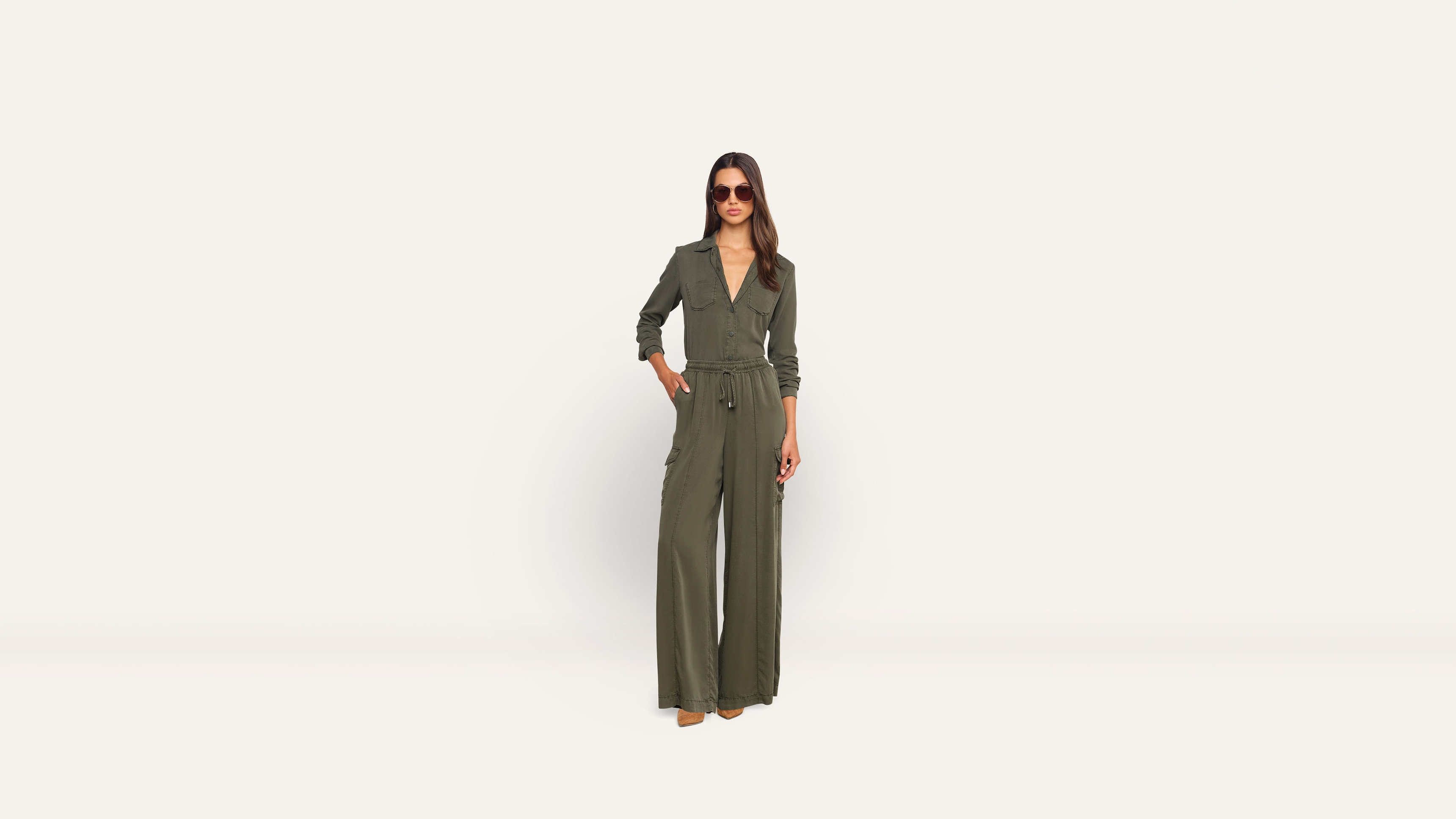 A woman wearing a long-sleeved olive green jumpsuit and sunglasses poses against a plain background.